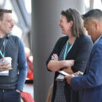 Three people standing chatting. One man is holding a coffee. Another is on his phone.